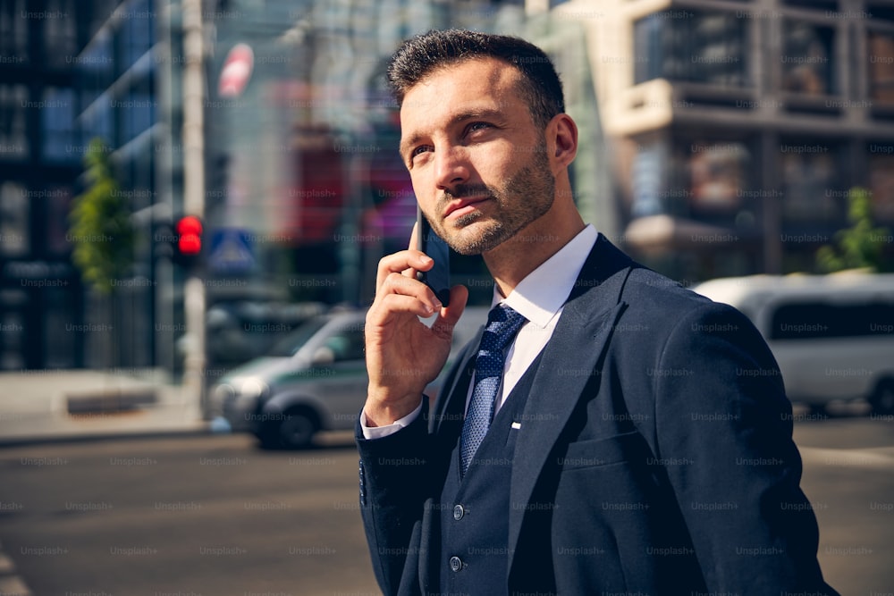 Bel homme debout sur l’arrière-plan du centre d’affaires moderne et parlant par téléphone pendant la promenade