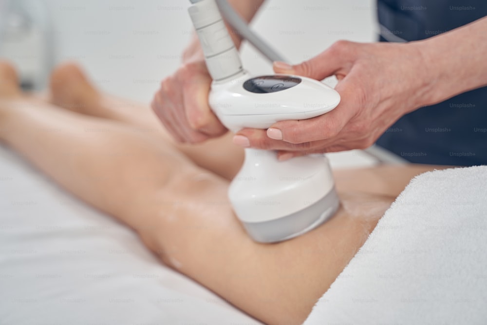 Hands of cosmetologist applying tool for ultrasound cavitation on the leg of woman during beauty treatment