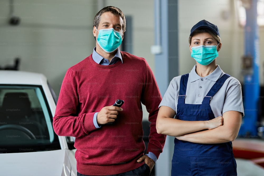 Auto repairwoman and her male customer with protective face masks standing in a workshop and looking at the camera.