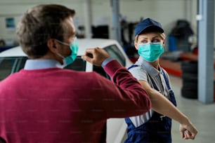Female car mechanic and her customer greeting with elbows while wearing protective face masks in auto repair shop during coronavirus epidemic.