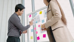 Business people work on project planning board in office and having conversation with coworker friend to analyze project development . They use sticky notes posted on glass wall to make it organized .