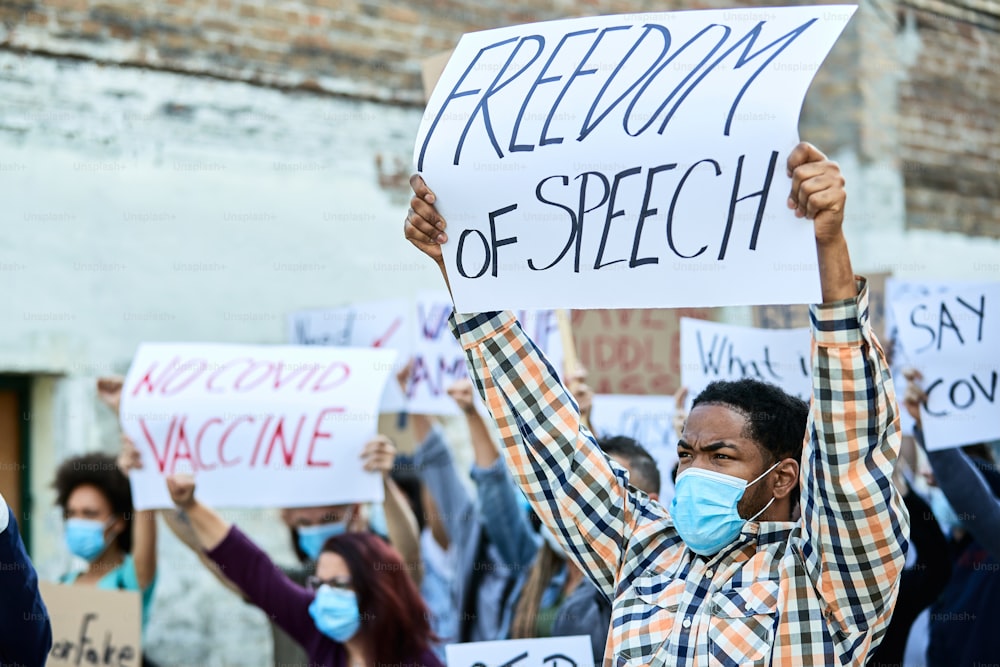 Große Gruppe unzufriedener Menschen, die während der Coronavirus-Pandemie protestieren. Der Fokus liegt auf einem schwarzen Mann, der ein Banner mit der Aufschrift "Redefreiheit" hält.