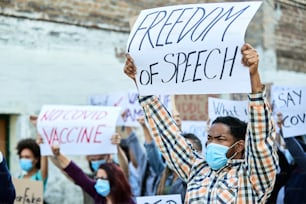Large group of displeased people protesting during coronavirus pandemic. Focus is on black man holding a banner with Freedom of speech inscription.