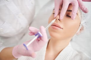 Skilled beautician holding client nose and performing injectable procedure with hyaluronic acid syringe