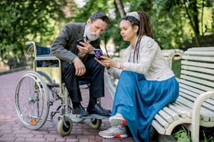 An elderly patient sitting in a wheelchair, talking with his carer, nurse, or granddaughter, sitting on the bench in park, looking at their smartphones, smiling and having fun.