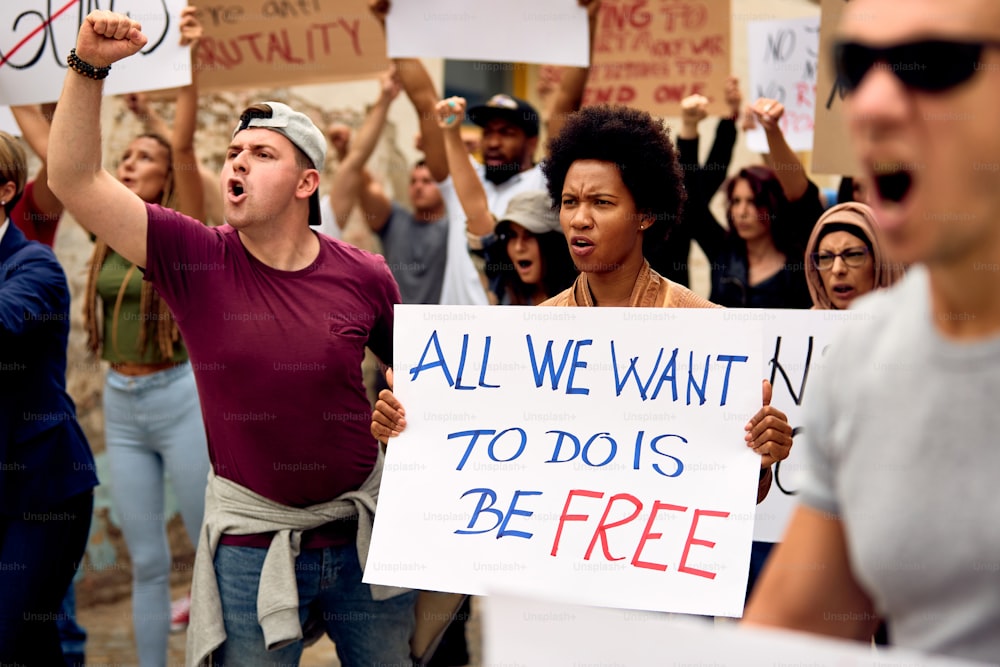 Displeased multi-ethnic crowd of people protesting for human rights on city streets. Focus is on black woman holding placard with freedom inscription.