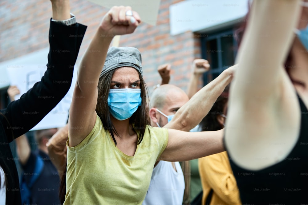 Female activist wearing protective face mask while protesting with crowd of people during COVID-19 pandemic.
