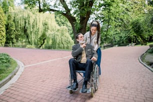 향취를 가진 행복한 젊은 힙스터 현대 여성, 그녀의 늙은 선배 할아버지와 함께 걷고, 휠체어에 앉아 공원의 야외에서 이야기하고 웃고 있습니다. 간병인과 함께 있는 장애인 남성.