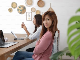 Portrait of female university student sitting on chair at table in cafe with her friend and digital devices