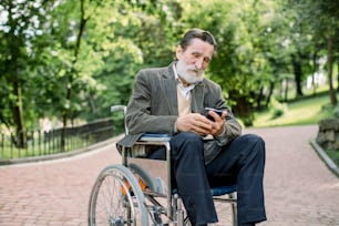 Bearded Caucasian old man is sitting in a wheelchair in the park alone, using his smartphone for serfing internet or socail networks.