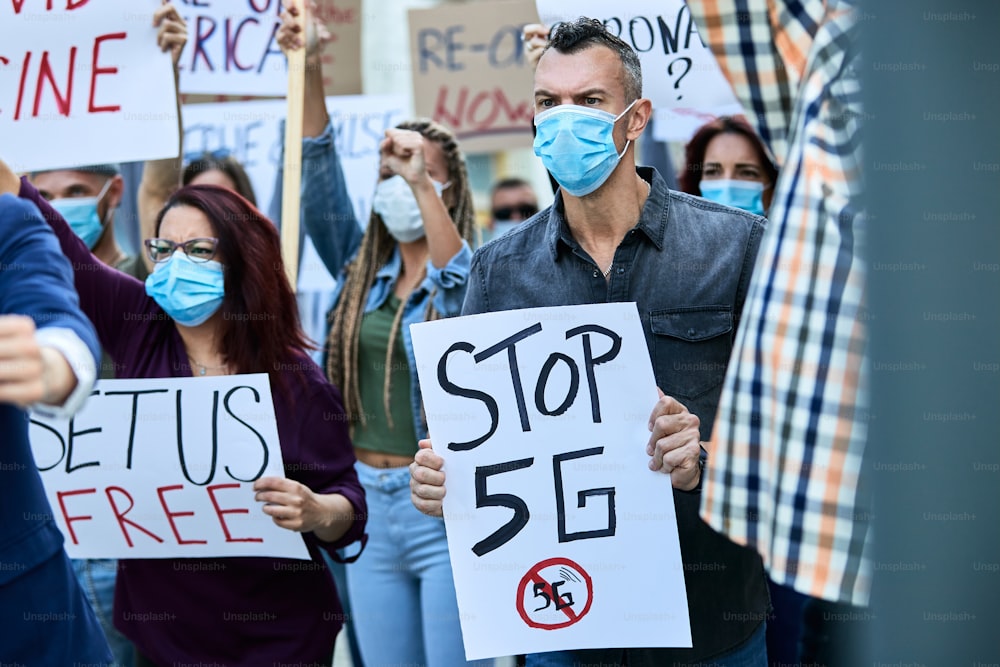 Young Caucasian man carrying placard with Stop 5G while protesting with crowd of people during coronavirus epidemic.