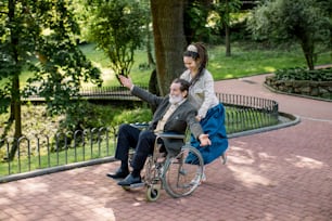 Care helper woman, pretty young hipster granddaughter walking with happy elderly disabled man with arms outstretched, pushing a wheelchair and running in the city park in summer day