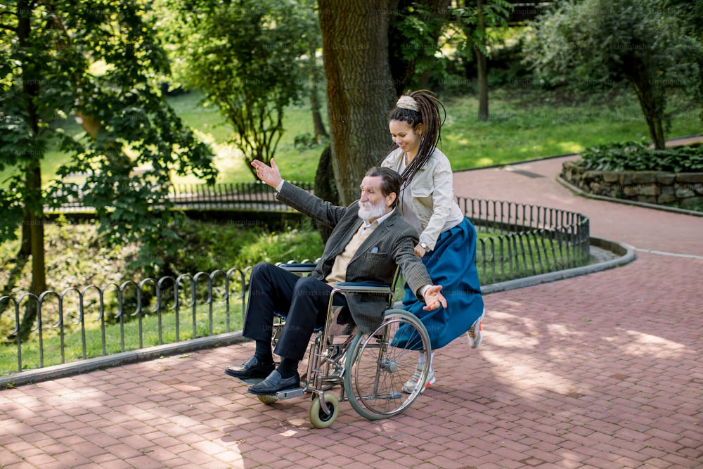 케어 도우미 여자, 예쁜 젊은 힙스터 손녀는 행복한 노인 장애인과 함께 팔을 뻗고, 휠체어를 밀고, 여름날 도시 공원에서 뛰고 있다