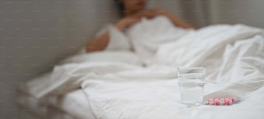 A glass of water and medicine are putting on the bed over an ill woman is a background.