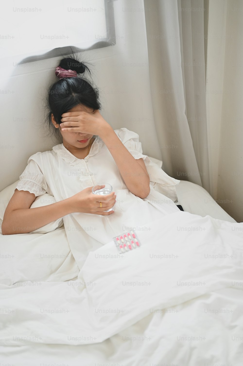 病気の女性がコップ一杯の水を持ち、寝室のベッドに横たわりながら錠剤を飲んでいます。