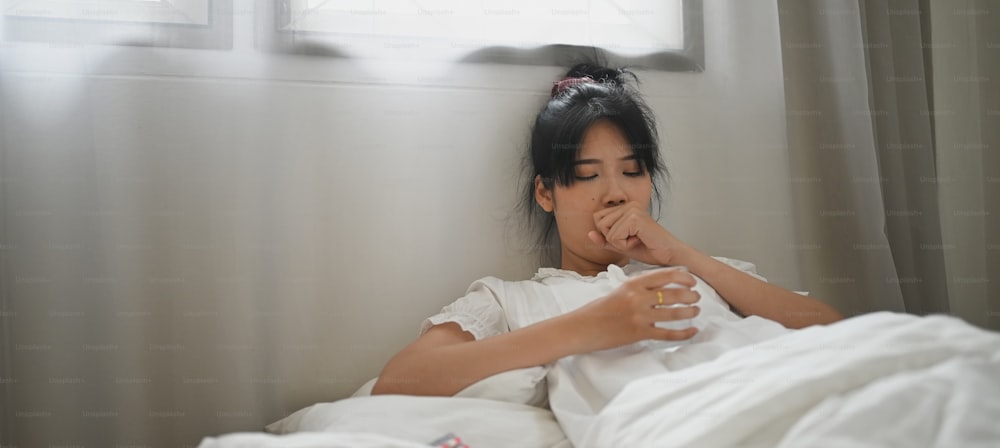 Una mujer enferma sostiene un vaso de agua y consume una pastilla mientras está acostada en la cama del dormitorio.