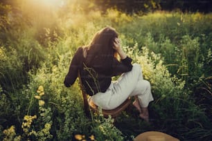 Stylish elegant girl sitting on rustic chair in sunset light in summer meadow. Fashionable woman relaxing in summer countryside. Creative image. Atmospheric tranquil moment