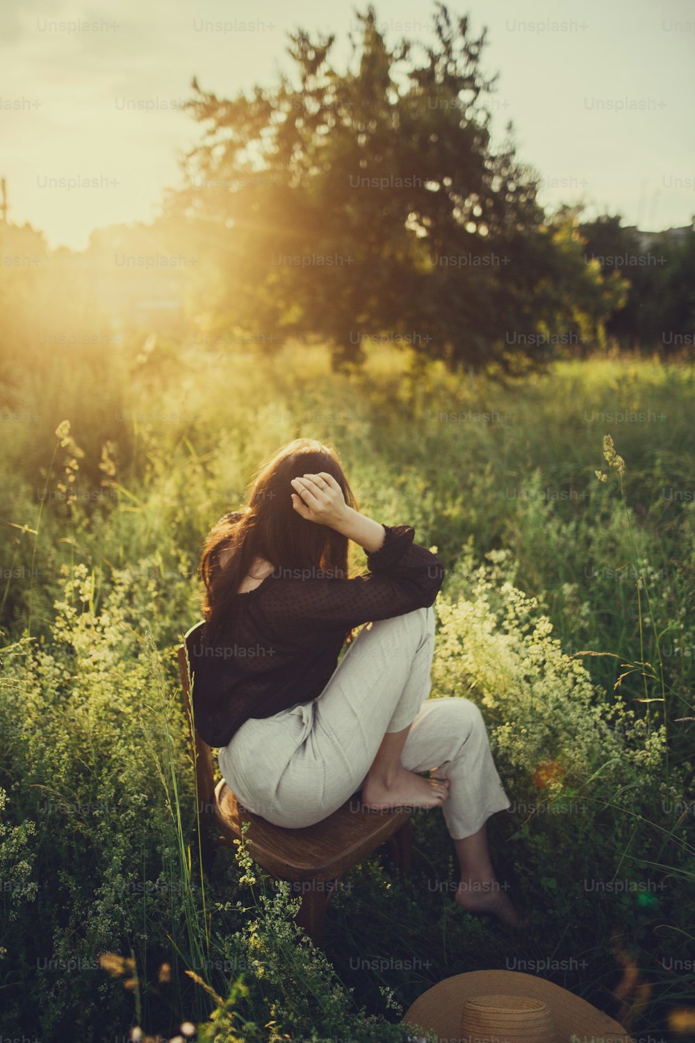 Slow living. Fashionable woman relaxing in summer countryside. Stylish elegant girl sitting on rustic chair in sunset light in summer meadow. Tranquil moment. Creative image.