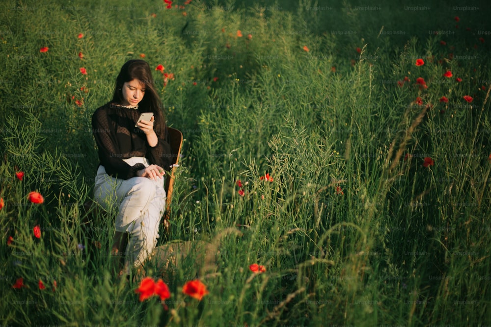 Fashionable young woman using smartphone making photos. Stylish elegant girl chatting online, holding phone and sitting in summer meadow in warm sunset light.