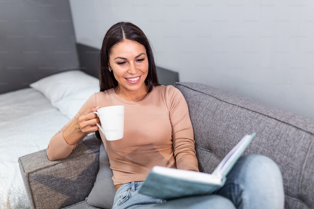 Happy young beautiful woman relaxing at home in cozy winter or autumn weekend with book and cup of hot tea or coffee, sitting in sofa and reading a book