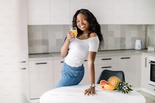 Happy mixed race dark skinned woman holding glass with fresh juice, looking to camera, while leaning at the table with fresh fruits at modern home kitchen.
