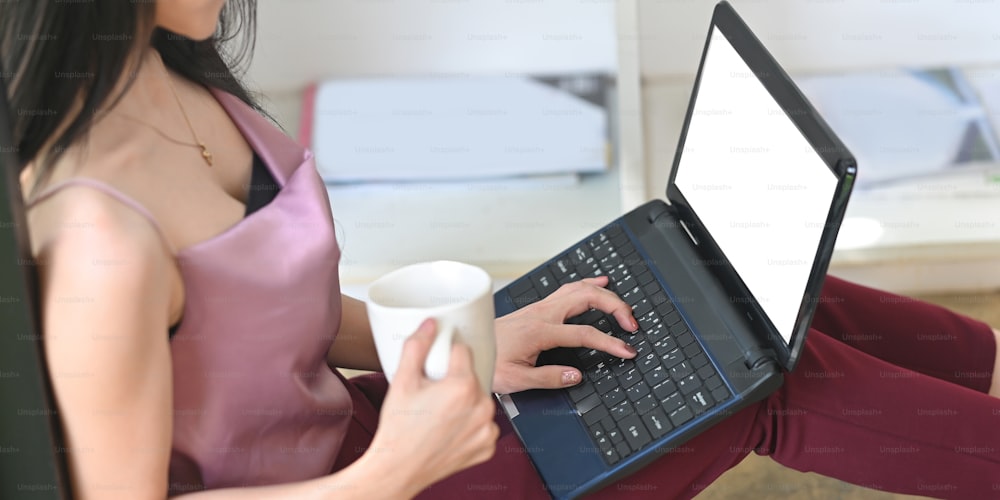Cropped image woman is drinking coffee and using a mockup computer laptop.