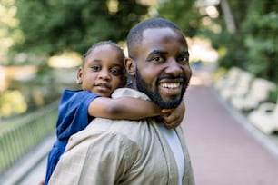 Smiling handsome African American father carrying his cute little daughter on back outdoors in city park. Daddy carrying her daughter on piggyback in the nature