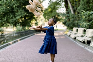 Portrait of happy little African girl child in green city park throwing up teddy bear toy, flying in the air. Smiling dark skinned kid girl in blue dress playing in summer park