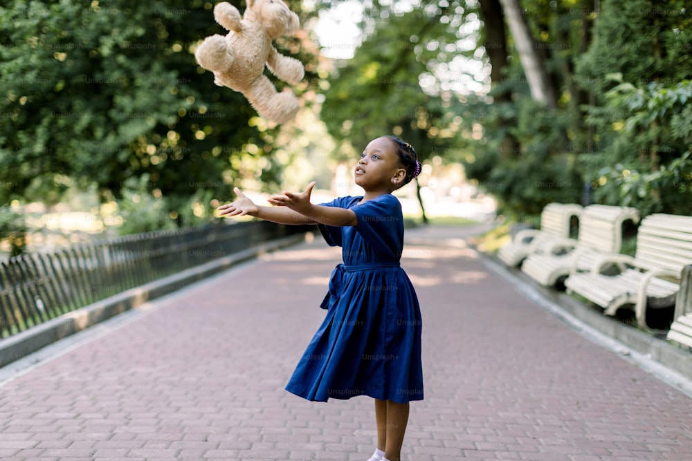 녹색 도시 공원에서 테디 베어 장난감을 던지고 공중에서 날아가는 행복한 작은 아프리카 소녀의 초상화. 여름 공원에서 놀고 있는 파란 드레스를 입은 웃는 어두운 피부의 어린 소녀