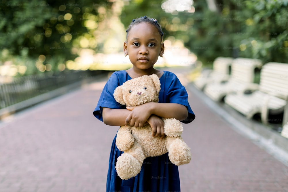 Outdoor portrait of little pretty African-American girl in stylish blue dress, smiling and holding teddy bear toy, looking at camera. Happy childhood concept.