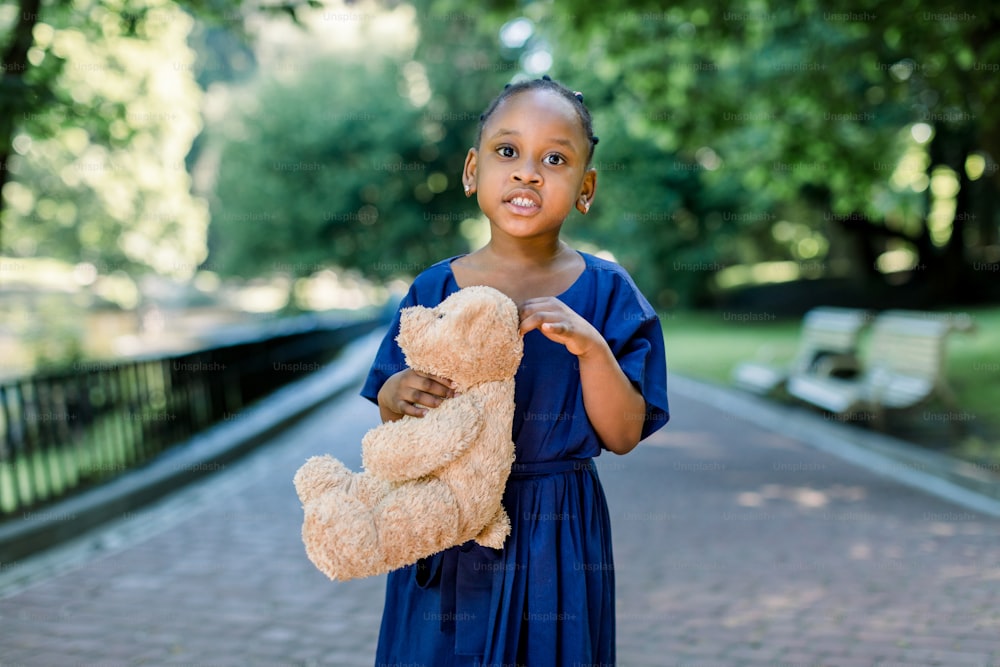 Pretty little African child girl in fashionable blue dress holding teddy bear toy while walking in nature park outdoors.