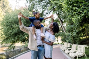 Young happy laughing African couple parents, spending time together and enjoying warm summer evening in park, walking with their little adorable daughter, sitting on shoulders of father.