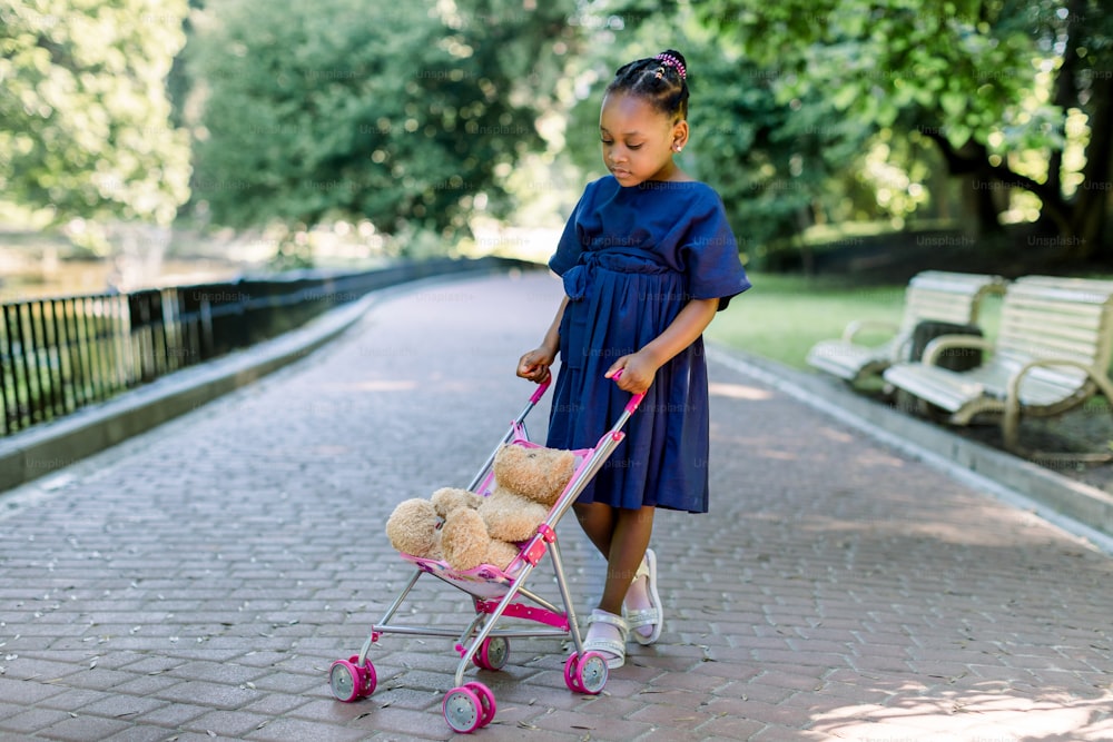 Beautiful little African girl in blue dress is playing with teddy bear toy, sitting in small kid stroller, walking in the beautiful city park. Outdoors summer portrait.