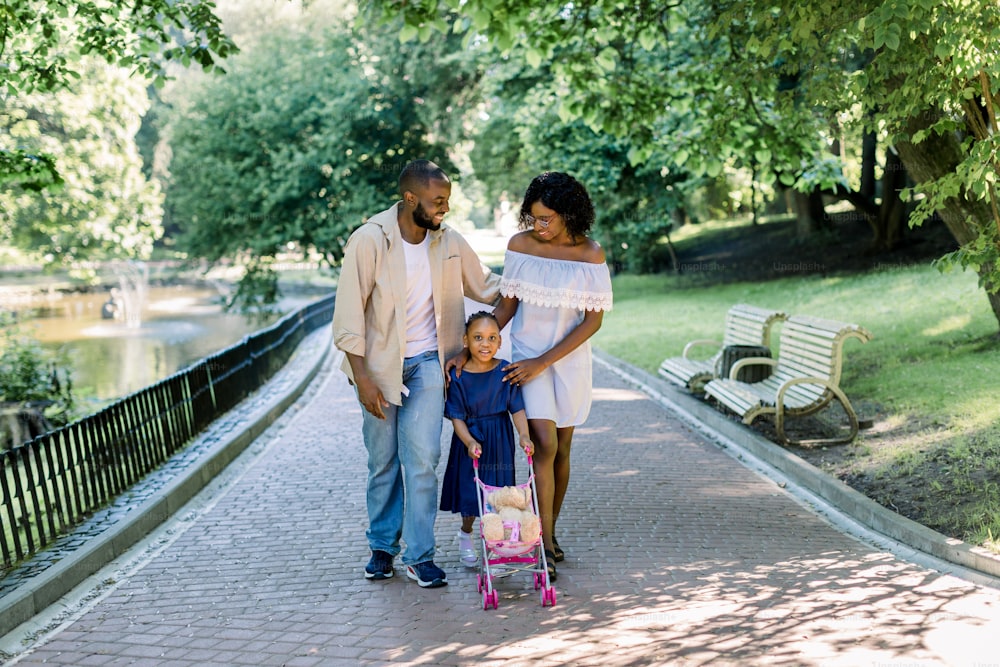 Giovane famiglia africana con la figlia piccola che si diverte nel parco della città naturale, camminando lungo il viale del parco. Bambina carina in vestito blu che gioca con il giocattolo del passeggino e l'orsacchiotto.