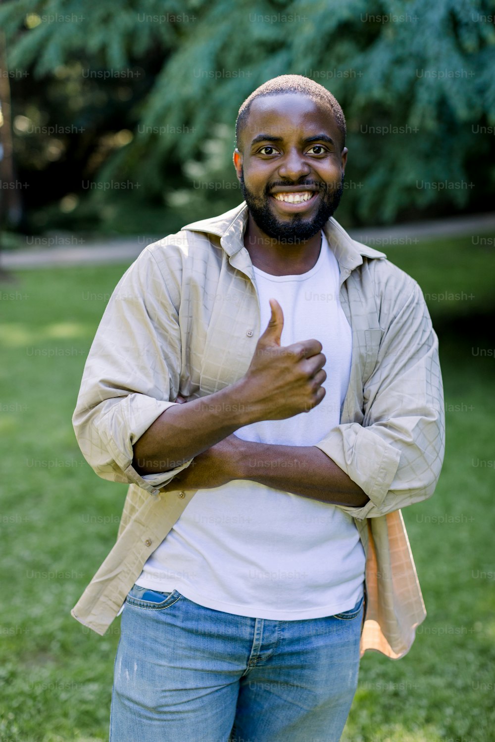 Outdoor casual lifestyle portrait of young African dark skinned man, wearing white t-shirt, beige shirt and jeans, smiling to camera in park nature, showing his thumb up.