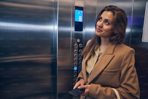 Cute brunette female in trendy coat staying in lift alone with mobile in hands and smiling
