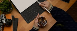 Toma aérea de la mano femenina sosteniendo una taza de café helado mientras trabaja con una tableta digital de maqueta en una mesa de madera