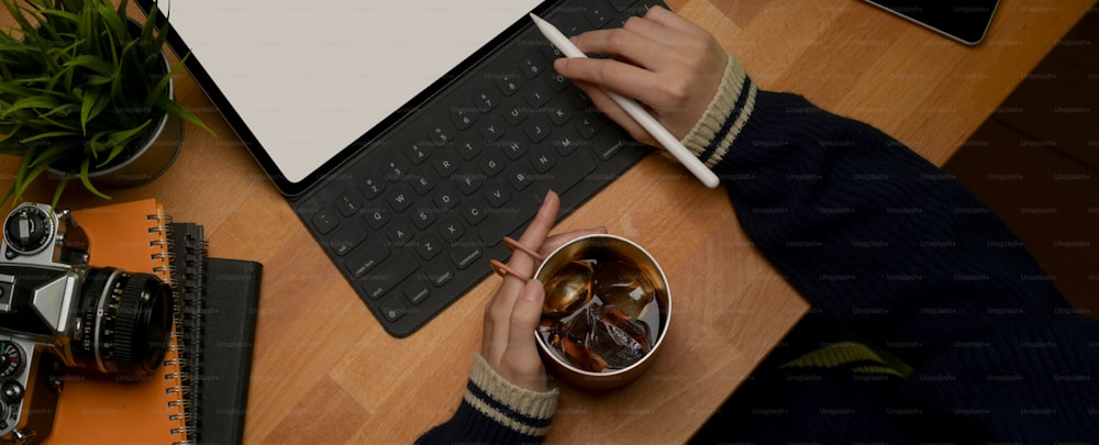 Overhead shot of female hand holding ice coffee mug while working with mock up digital tablet on wooden table