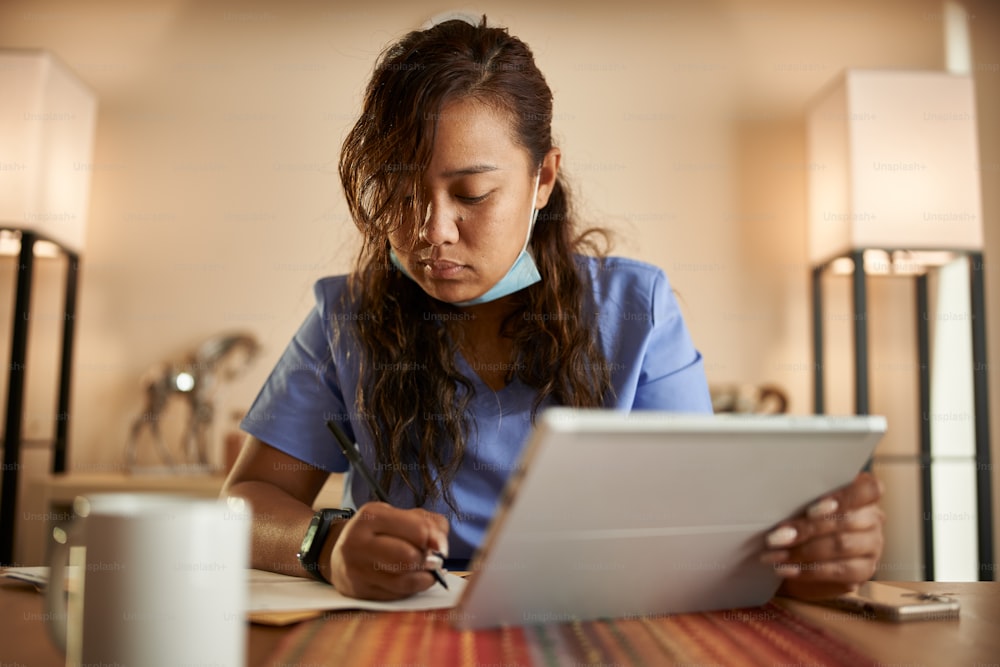 filipina nurse working from home doing paperwork and using tablet on table