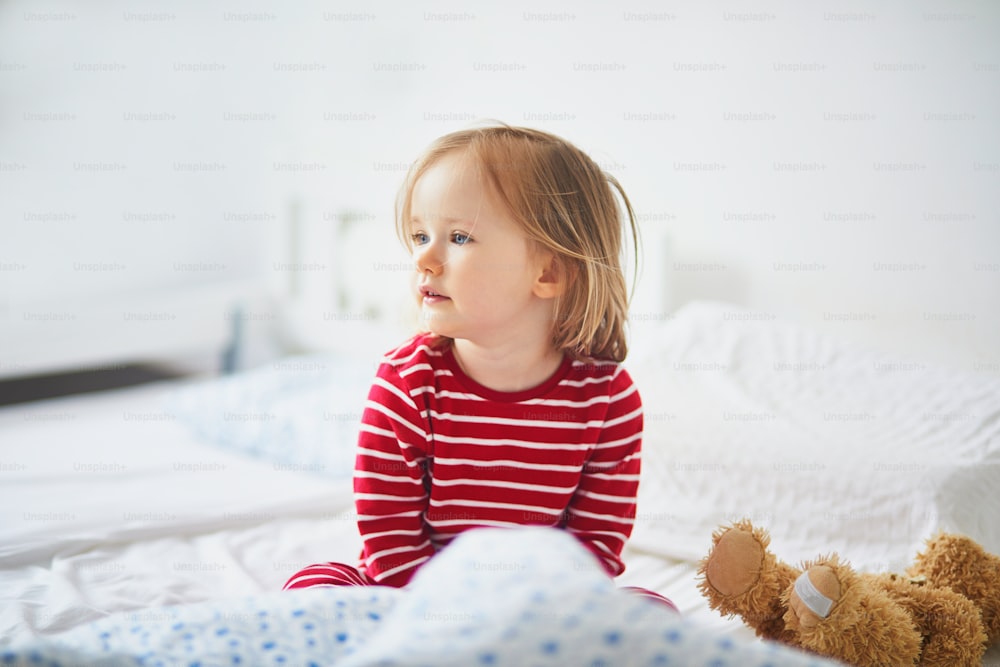 Happy toddler girl in striped red and white pajamas sitting on bed right after awaking. Day naps for small kids