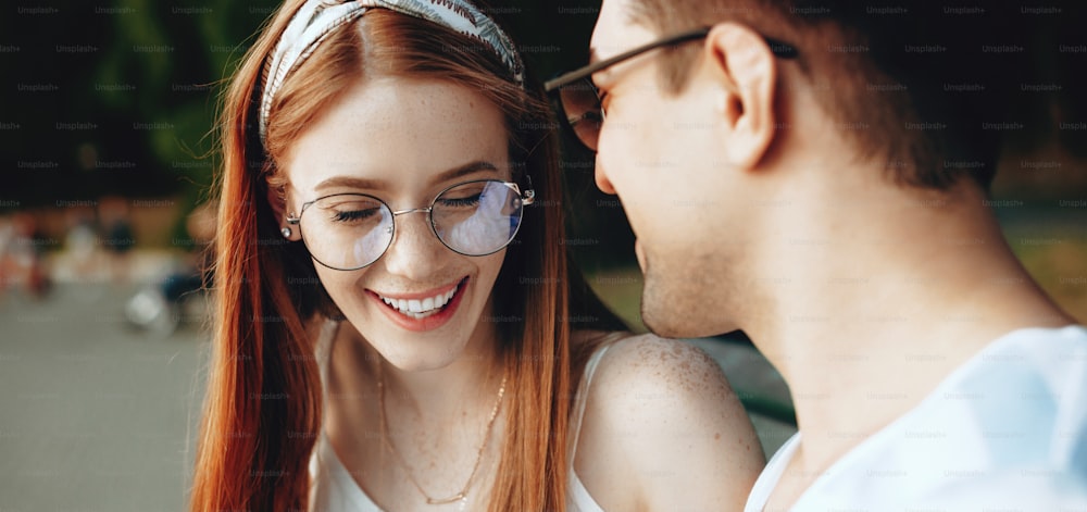 Red haired caucasian woman with freckles and eyeglasses laughing with her lover on the bench in the park