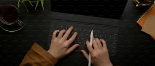 Overhead shot of female hands typing on tablet keyboard on dark office desk with schedule book and decoration