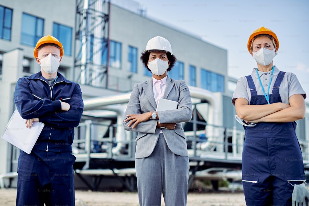 Afroamerikanischer Bauunternehmer und zwei Ingenieure tragen Schutzmasken, während sie mit verschränkten Armen auf der Baustelle stehen.