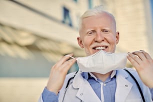 Happy albino doctor putting on protective face mask while looking at camera outdoors.