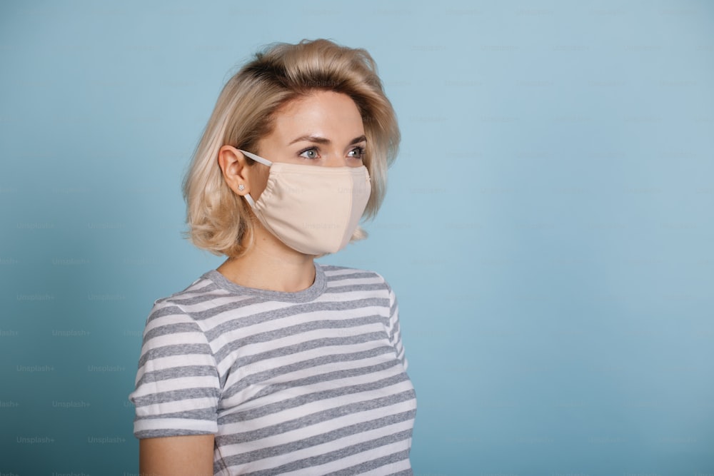 Side view photo of a caucasian blonde woman with a medical mask on face posing on a blue background with free space