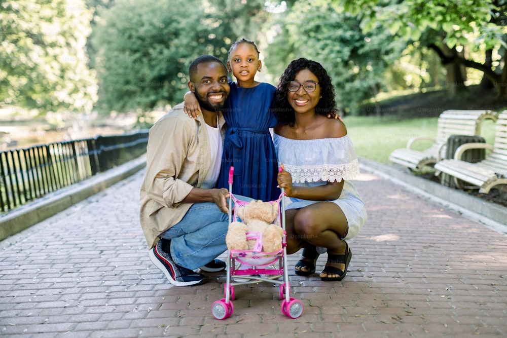 Lovely African family with small daughter, wearing stylish casual clothes and dress, standing outdoors in summer sunny park, laughing, playing together with kid stroller and teddy bear.