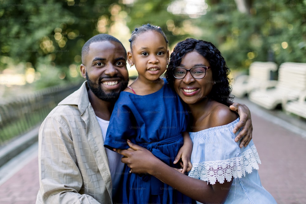 Close up portrait of cheerful young African couple with their child daughter, posing to camera with happy smiles, while enjoying their time together in park.