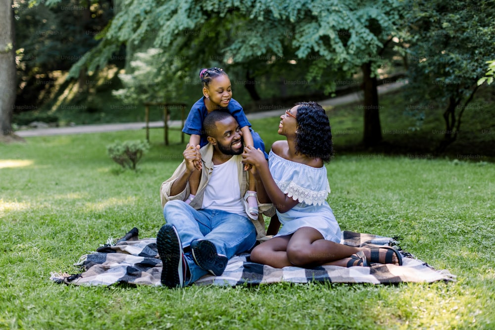 夏の日に公園でピクニックをする3人の幸せなアフリカ人家族。公園の屋外で、市松模様の毛布の上に座っている若い家族。子供の女の子はパパに便乗しています。