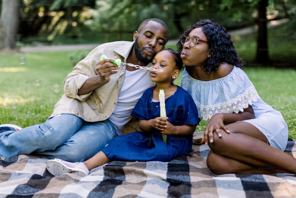 Familienzeit, Urlaub, Freizeit zu zweit. Fröhliche afroamerikanische Familie mit einem kleinen Kind, das zusammen Seifenblasen bläst, Spaß bei einem Picknick im Stadtpark hat und auf einer karierten Decke sitzt.