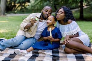 Family time, vacations, leisure together. Joyful African American famile with little kid girl blowing bubbles together, having fun on a picnic at city park, sitting on checkered blanket.